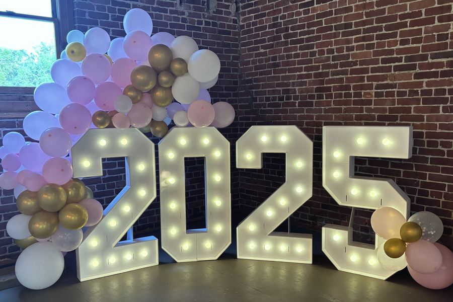 Lit marquee numbers 2025 against a brick wall, with a blush, white and gold balloon garland climbing one side.