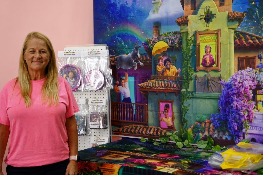 Sherri Purcell in her shop, beside a rack of Sherri's Heavenly Balloons party plates and a printed party backdrop.